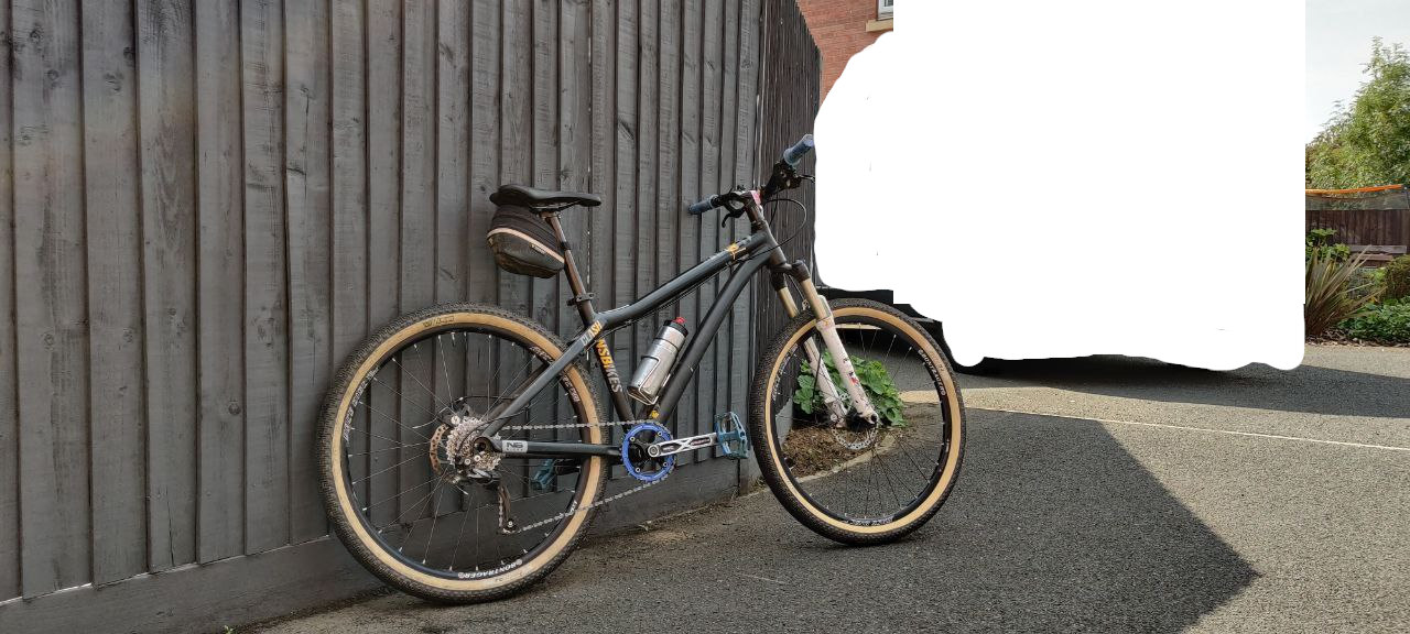 A view of the back of the bike, with tan wall tyres, blue chainring and pedals, black frame with yellow accents, and white fork. The cranks are black and silver