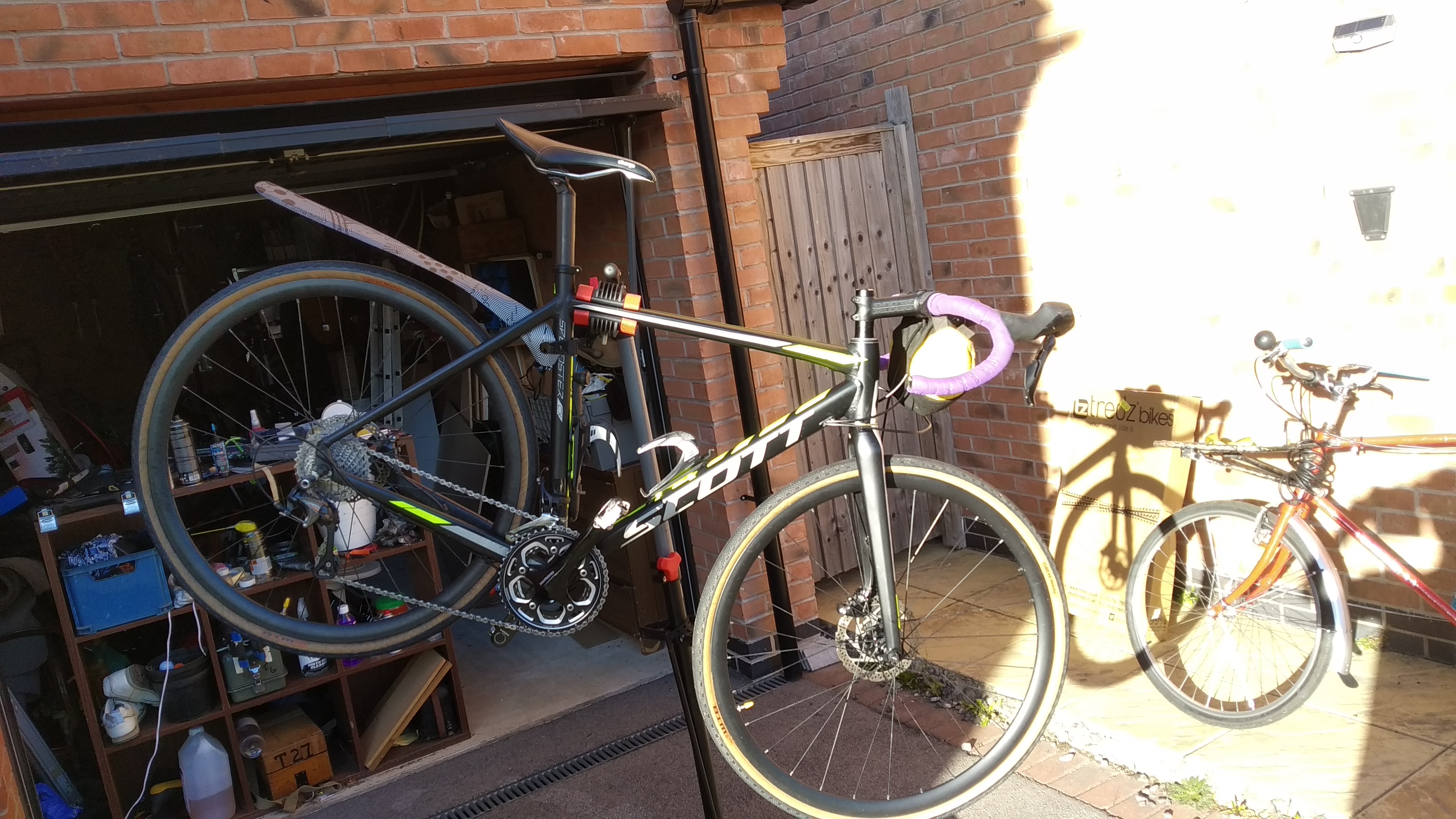 My CX bike in a stand. It's got a black frame with yellow and silver accents. The mudguard on the rear is a straight silver bit of plastic.