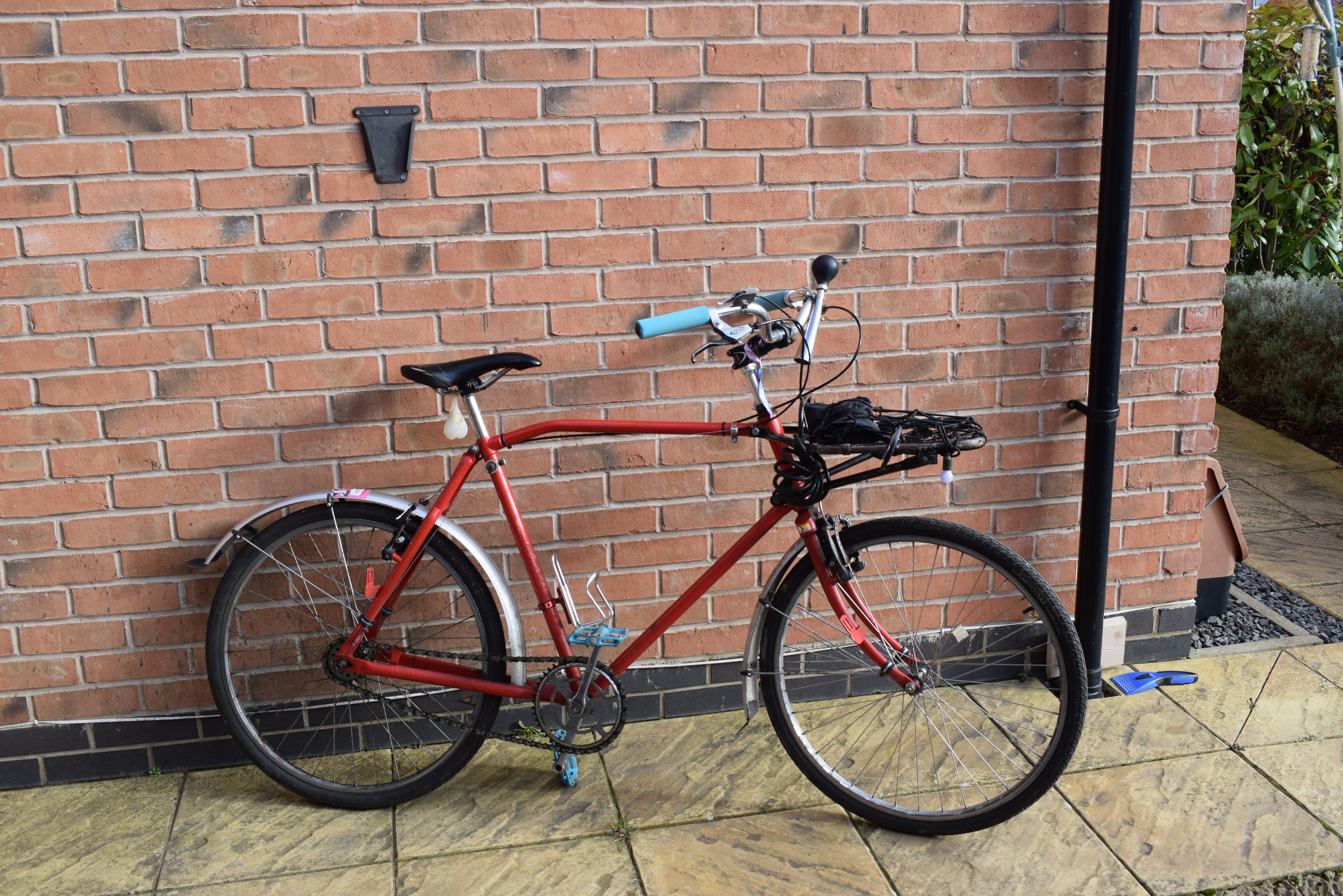 A side view of the bike, red, with light blue pedals and grips. A black saddle hangs a light that looks like a pair of bollocks. There are silver mudguards, spokes, and hubs, the chain sags slightly (as was the style at the time).