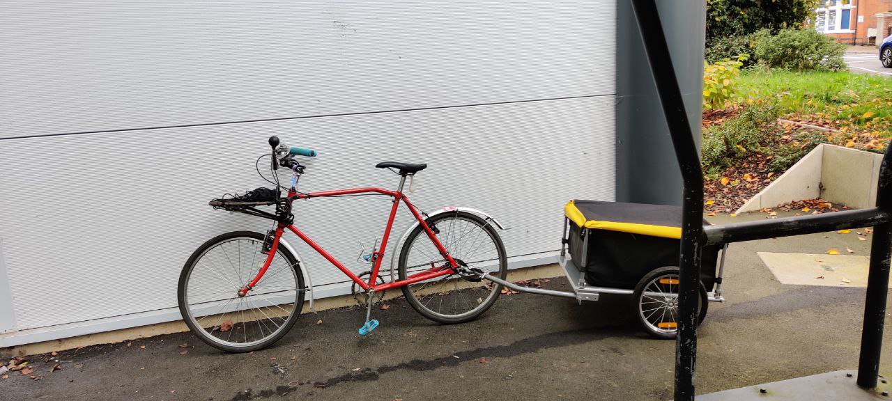 A red bike with a black, silver, and yellow accented trailer attached lent up against a wall outside a bike shed