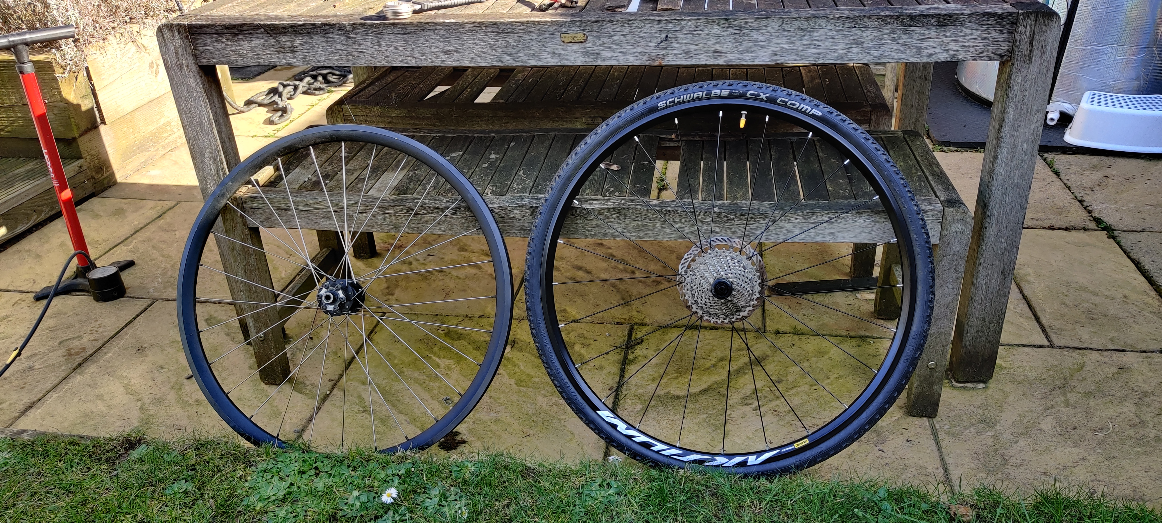 Two wheels next to each other. A black wheel with silver spokes and no tyre or cogs, and a shallower black wheel with a tyre and cogs with black spokes. It has white text on the black rim