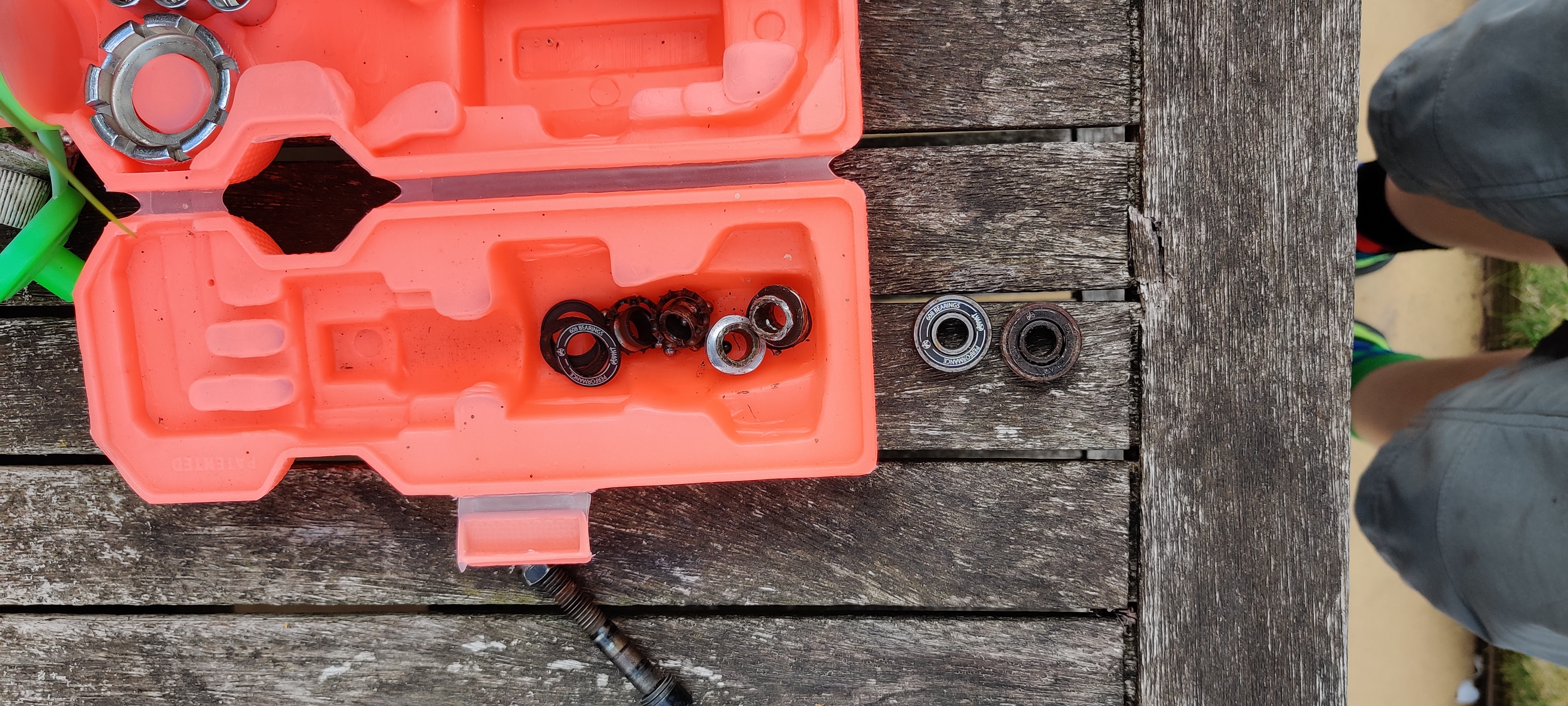 A waterbottle shaped toolkit in orange plastic with two destroyed but whole skateboard bearings below it on the table, and all the parts of two mangled and dismantled bearings inside it.