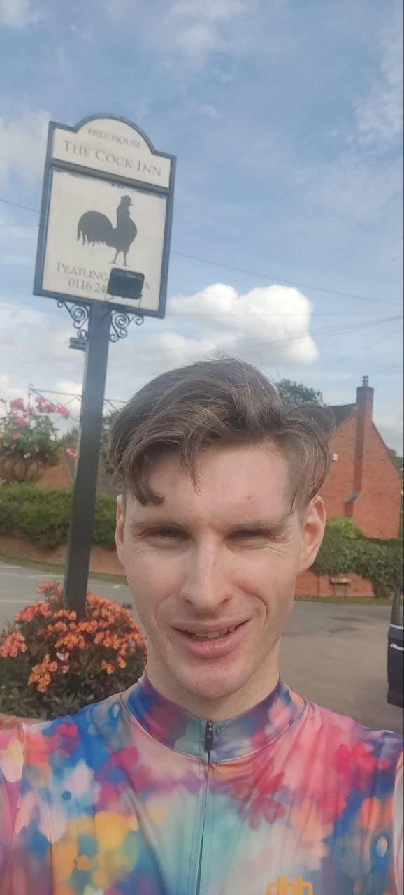 Me in a tie die floral jersey, smiling into the camera cheekily below a sign that says "The Cock Inn"
