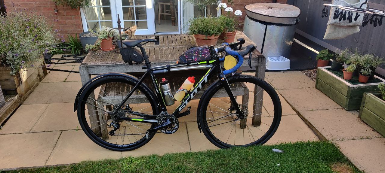 The fully set up bike in my garden. It's a black bike with yellow and white accents, blue bar tape, and 3 main bags on the saddle, top tube, and handlebars. There's a frame pump under the top tube, two large 750ml bottles, and a pride ribon on the stem and little flag on the fork crown.