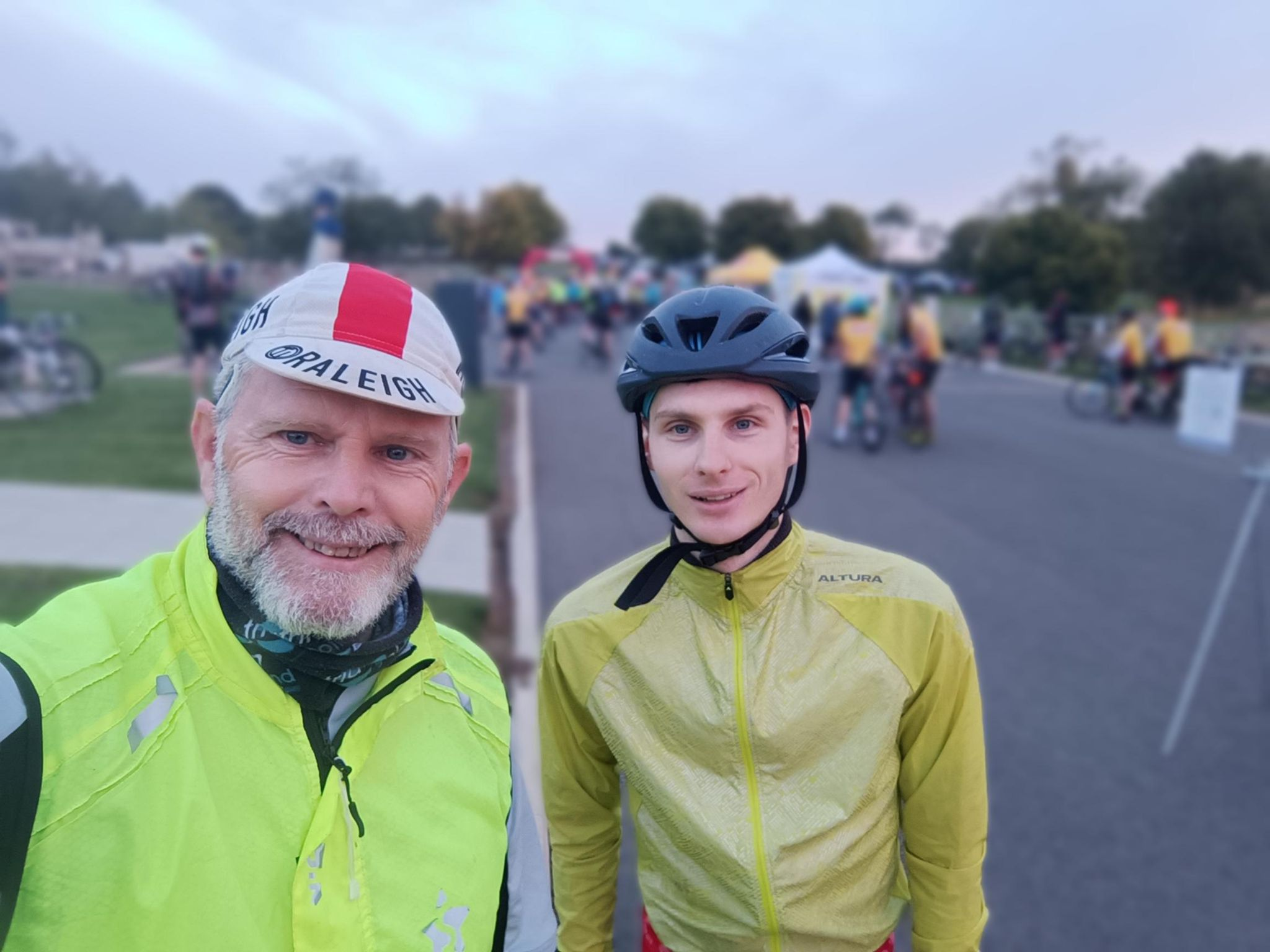 A photo of two people, me in a blue helmet and yellow jacket on the right of picture, and my Dad in a red and white Raleigh cap and yellow jacket on the left of picture at the start of the ride.
