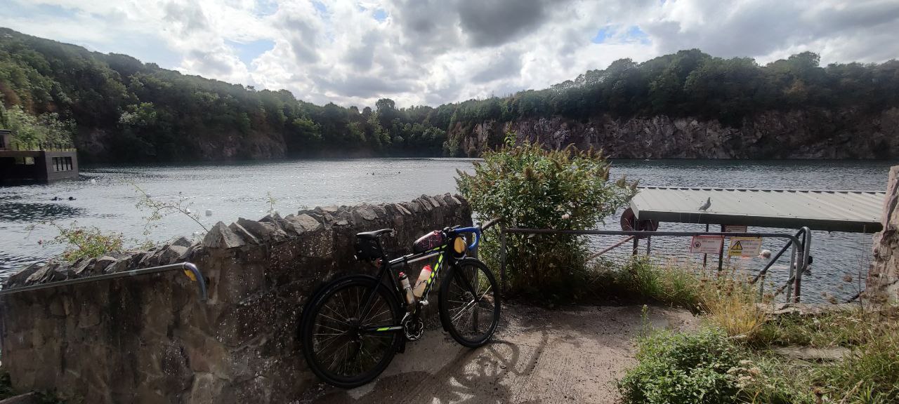 My bike overlooking the waters of Stoney Cove. It's lent up against a brick wall, there's a small tree in front, and the rock faces lead up into trees on the other side of the river