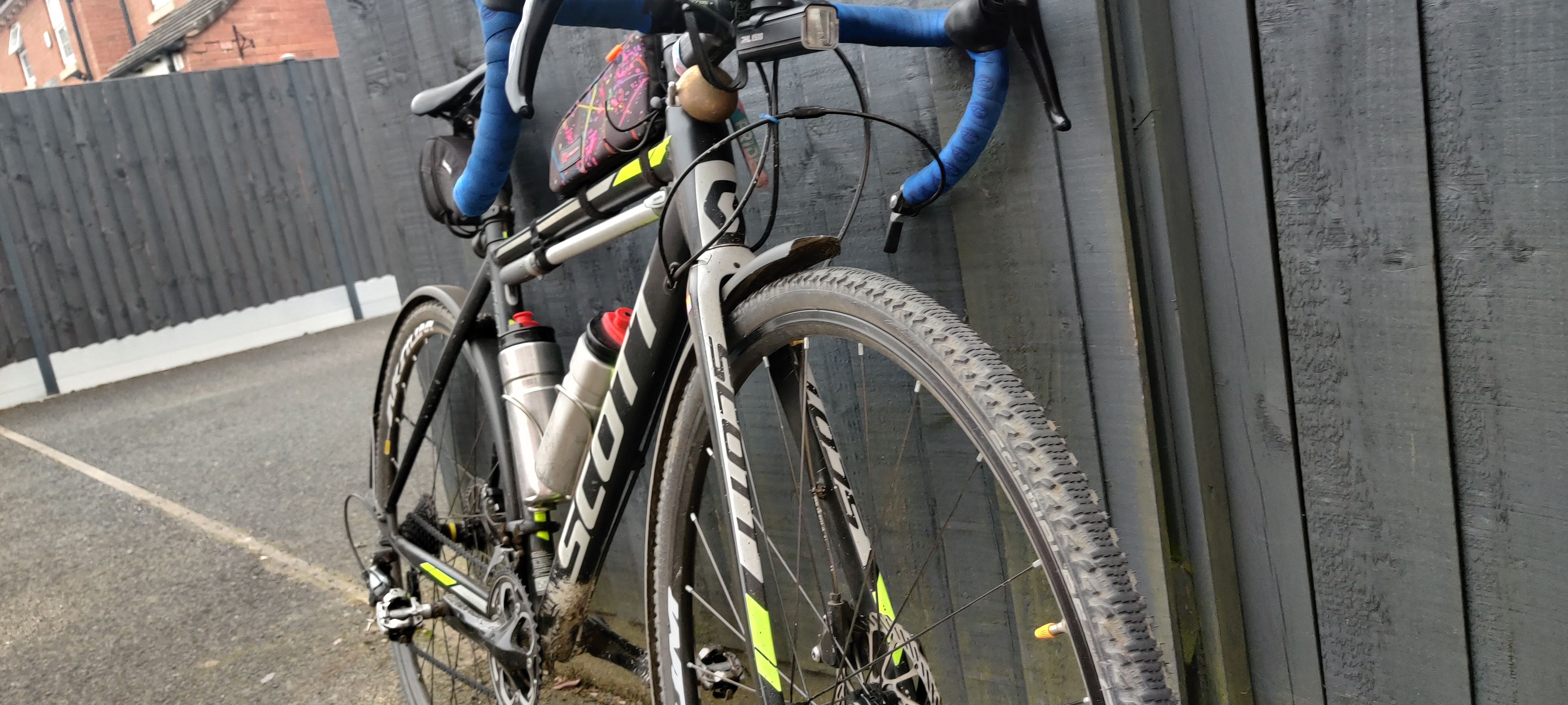 The front of my bike from a diagonal angle. It's mostly black with light grey parts, and yellow accents. The bar tape is blue and there's a swirly patterned bag on the top tube. Parts of the bike are covered in splatters of mud, especially around th tyres. The wheels and mudguards have a thin layer of dust on them, almost inperceptable in the picture because it's even all over The front of my bike from a diagonal angle. It's mostly black with light grey parts, and yellow accents. The bar tape is blue and there's a swirly patterned bag on the top tube. Parts of the bike are covered in splatters of mud, especially around th tyres. The wheels and mudguards have a thin layer of dust on them, almost inperceptable in the picture because it's even all over