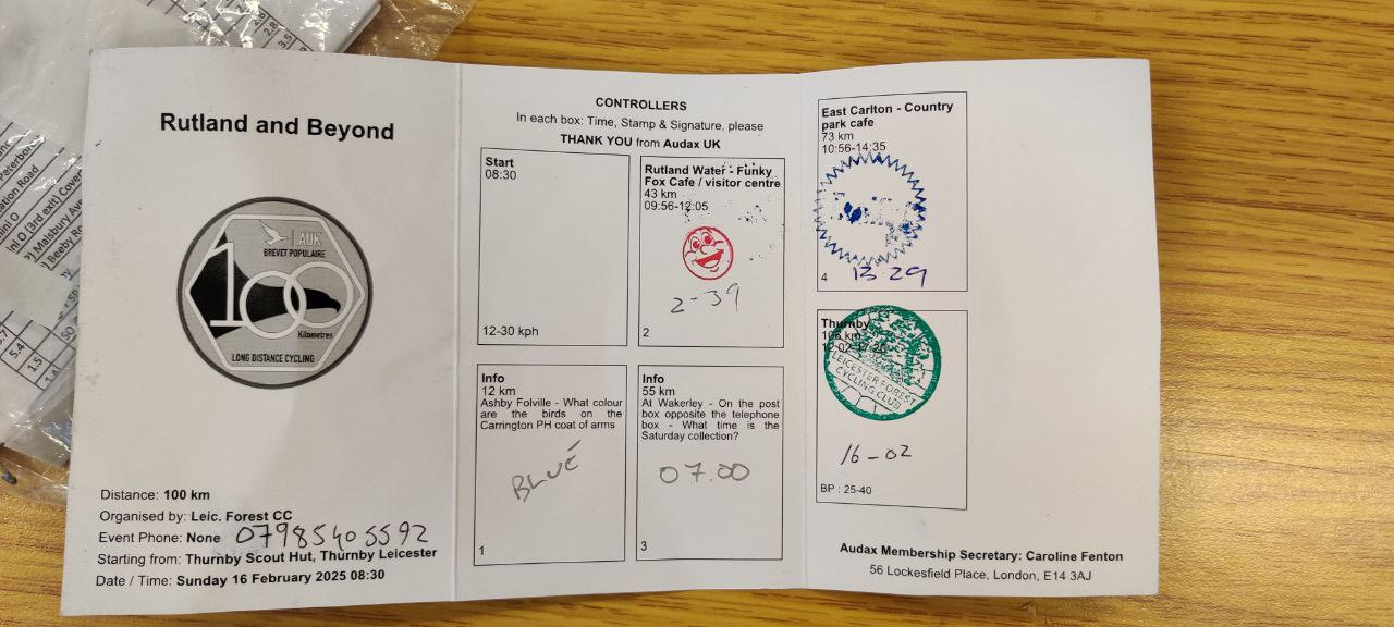 A card with 'Rutland and Beyond' and 100km audax stamp on the first third, then 4 boxes for first the start, then the first info answer of blue, then a stamped smiley face for the first cafe stop, the second info answer of 07:00, and finally across on the 3rd segment the two stamps for East Carltons second cafe stop, and the finish at 16:02.