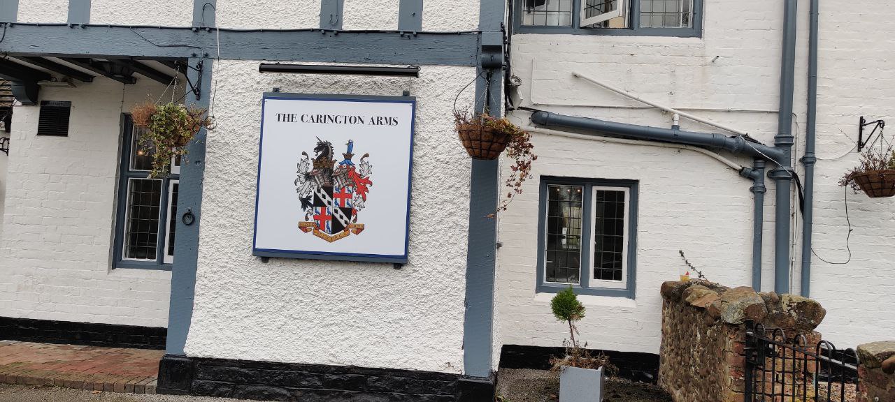 A photo of a white walled pub called 'The Carington Arms'. The crest is made up of a horse, a strange blue bird thing with something around it's neck, lots of feather like things, and the main shield is black in the upper left and lower right corners, with white stripe, and black diamonds diagonally across it. Then the other two corners are St Georges Cross, white background, red plus, with blue peacocks in each segment.