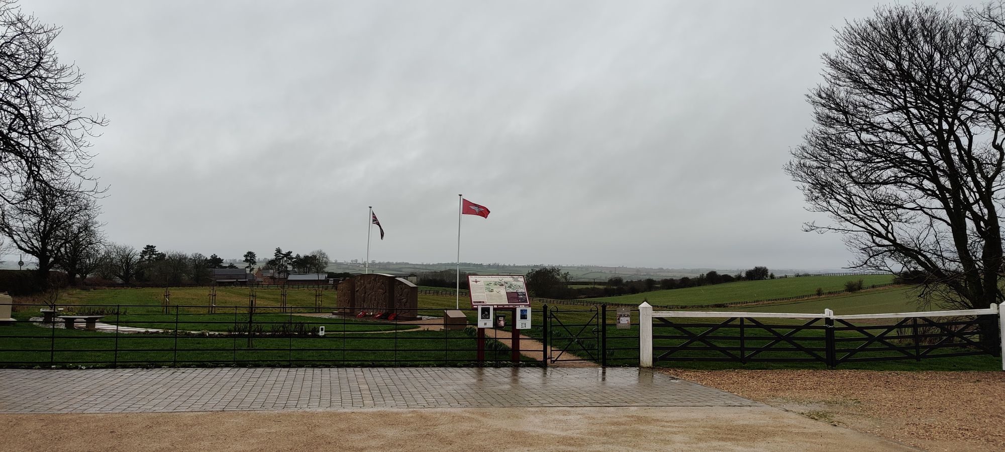 A view over fields at a war memorial with two flags blowing in the wind. There's a concrete memorial, a few benches, and an information sign board. The weather looks bad and wet and cold