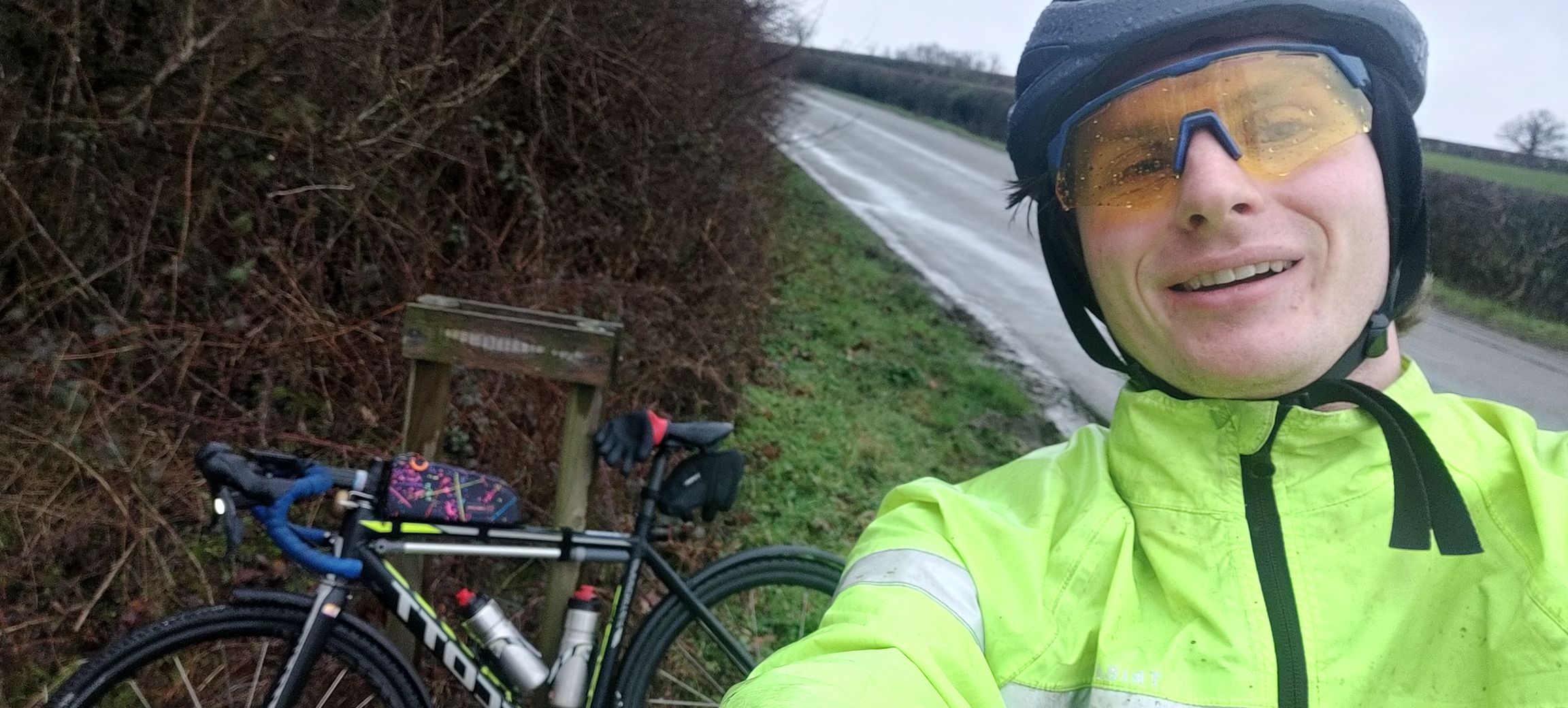 A picture of me in a yellow coat and yellow glasses, soaking wet, forcing a smile taking a picture with the "Knotting Hill" sign my bike is lent against