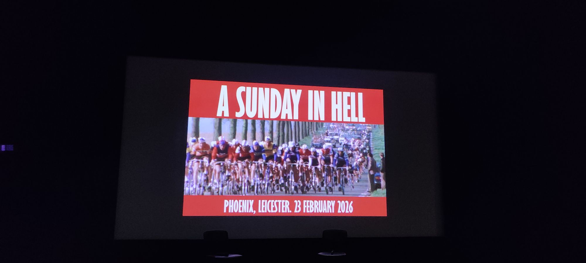 A cinema screen showing the image of a peloton of cyclists in the middle of the screen, brightly coloured in their 70s kit. The top is bold font A SUNDAY IN HELL, and the bottom "Phoenix, Leicester, 23 Februrary 2026" in white on a red background letterboxing the cyclists