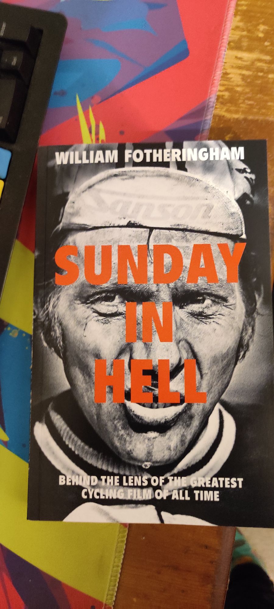 A copy of the book "Sunday in Hell" by William Fotheringham. The cover is of a dirty cyclist in black and white grimacing at the camera as water drips off his chin. The subtitle below is "Behind the lens of the greatest cycling film of all time" and the title "Sunday in Hell" is stamped in all caps and bright red across the face of the man